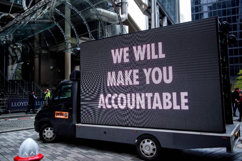 a black van with a digital display board on the back reads 'WE WILL MAKE YOU ACCOUNTABLE' in large pink letters. The ban is parked outside the front of Lloyds of London, a building with many insurance companies within it.