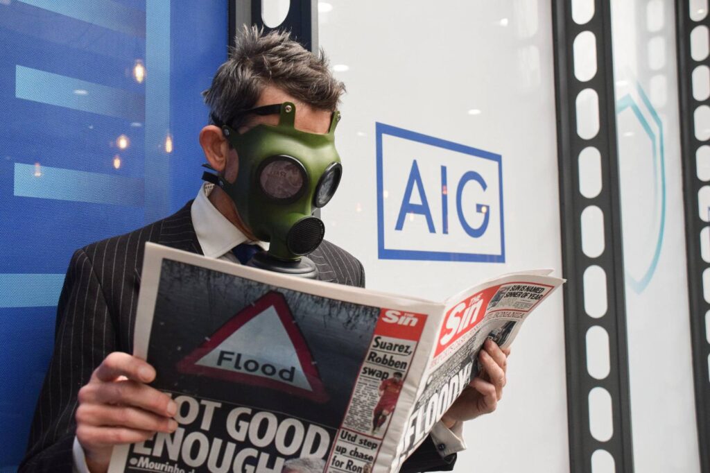 a protester stands outside the building owned by AIG insurers. The AIG logo can be seen clearly. The protester wears a gas mask while reading a newspaper with headlines representing climate chaos. The headline 'FLOODY HELL' can be seen, and a traffic warning sign for 'flood' is on the back page.