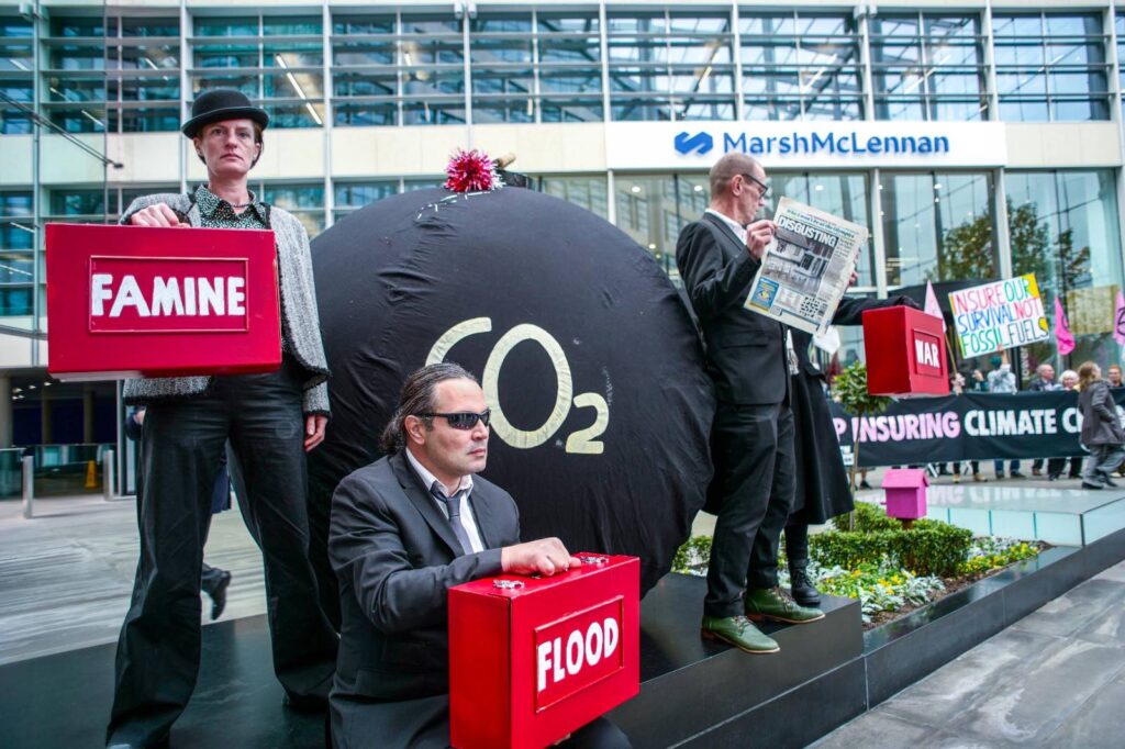 In the foreground, 3 Activists dressed professionally stand defiantly on a raised seating area outside the glass-fronted building of the MarshMcLennan Insurers head office, holding up red 'budget boxes' with the words 'FAMINE', 'FLOOD' and 'WAR'. A large black, round inflatable prop (about 2 metres tall) with white letters 'CO2' on it to represent a "carbon bomb" is behind them. In the distance, a large banner can be seen 'INSURING CLIMATE CHAOS' and with more protestors behind it.