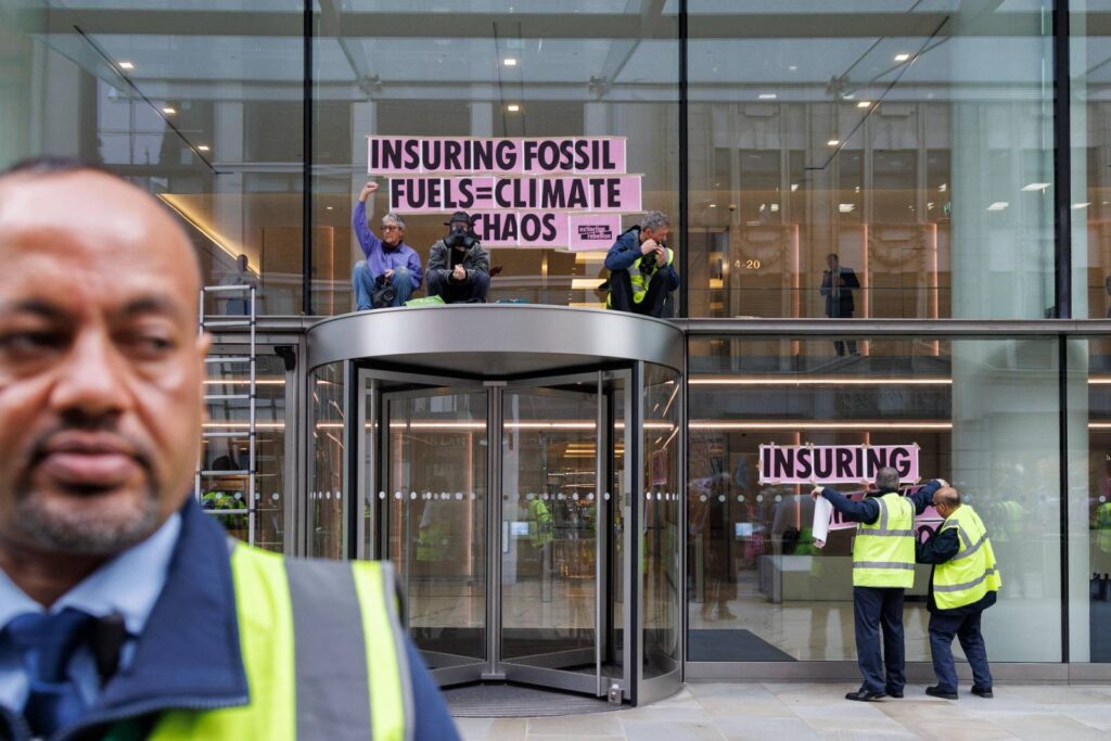3 protesters - one with an arm raised in a fist of defiance - sit on top of the revolving door canopy at the Walkie Talkie building in London. Large letters printed on pink paper spelling out 'INSURING FOSSIL FUELS = CLIMATE CHAOS' are temporarily fixed to the glass front of the building. 2 people in high-vis at the ground floor are adding more lettering to the front. The word 'INSURANCE' can be read. In the near focus, a member of security looks past the camera